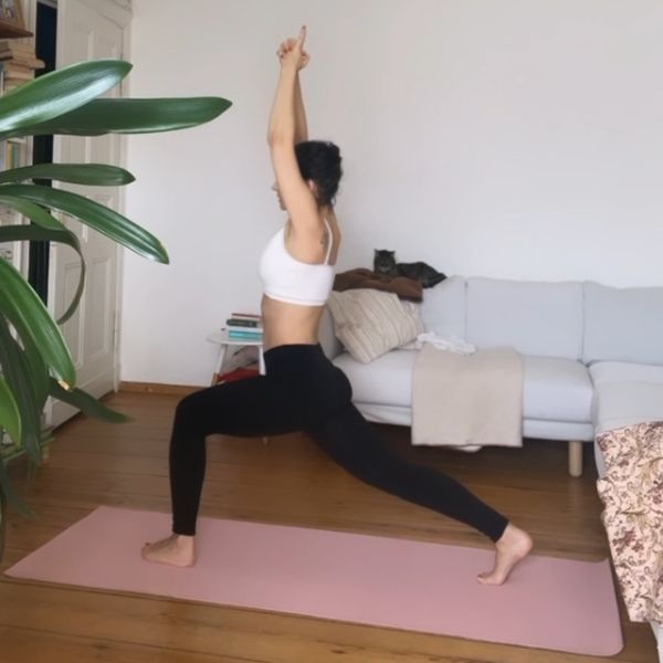 Woman practicing Yoga in her living room on a pink yoga mat. Warrior Yoga Pose. Her cat sits on the coach and is watching her.