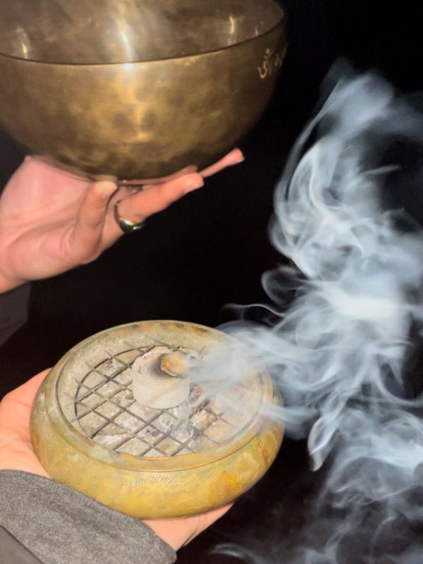 Dark background: Two hands, in one is a singing bowl, the other hand holds an incense burner, burning incense