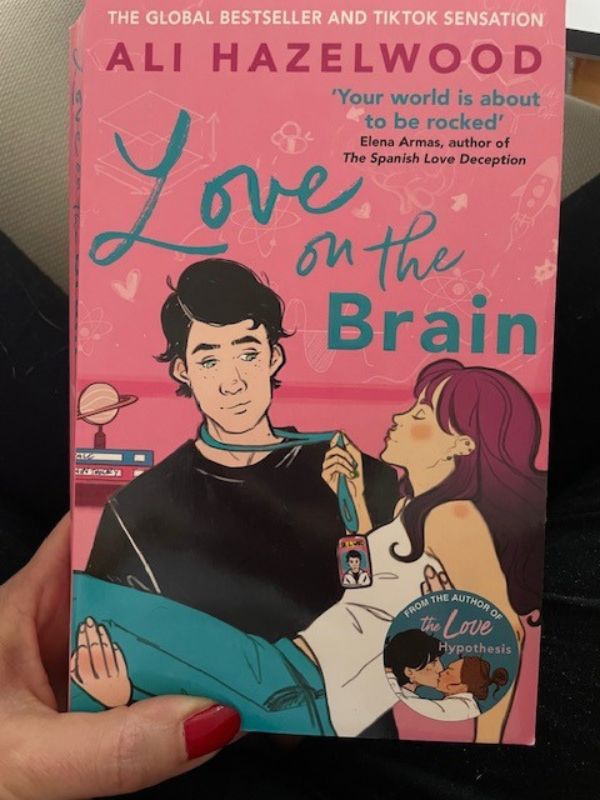 The book cover of "Love on the Brain" by Ali Hazelwood is a beautiful artwork that shows two people a man and a woman. The man is carrying the woman in his arms looking a bit confused or even scared. The woman seems to be unconscious. She has purple hair. 
The background of this book cover is pink, the title of the book is in blue. 