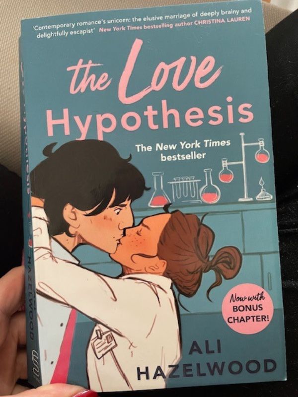 Book by Ali Hazelwood titled "The Love Hypothesis", the cover is in turquoise and shows two scientists in a lab kissing. The title is in pink.