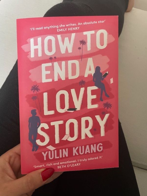 Female hand holds a book by Yulin Kuang with the title: 'How to end a love story', the color of the book is covered by pink-red shades. 