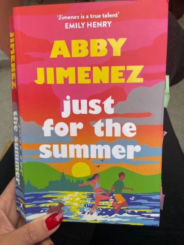 Hand holds a book by Abby Jimenez with the title: 'Just for the summer'; the book cover is a beautiful artwork showing a couple happily running in the low water of a lake, followed by a dog; the sun is setting behind the mountains.