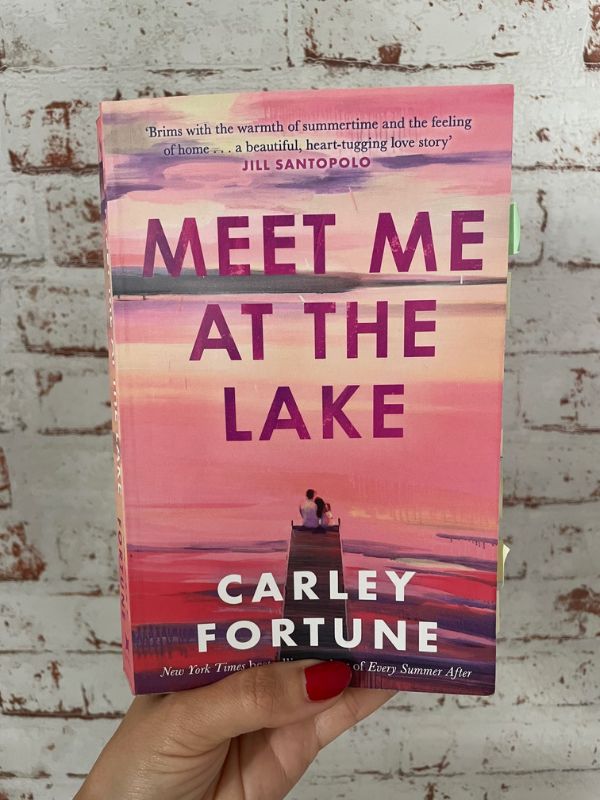 A hand is holding up a book in pink colored shades titled: 'Meet Me at the Lake' by the author Carley Fortune. It further shows a couple, sitting on the dock at a lake watching the sunrise. 