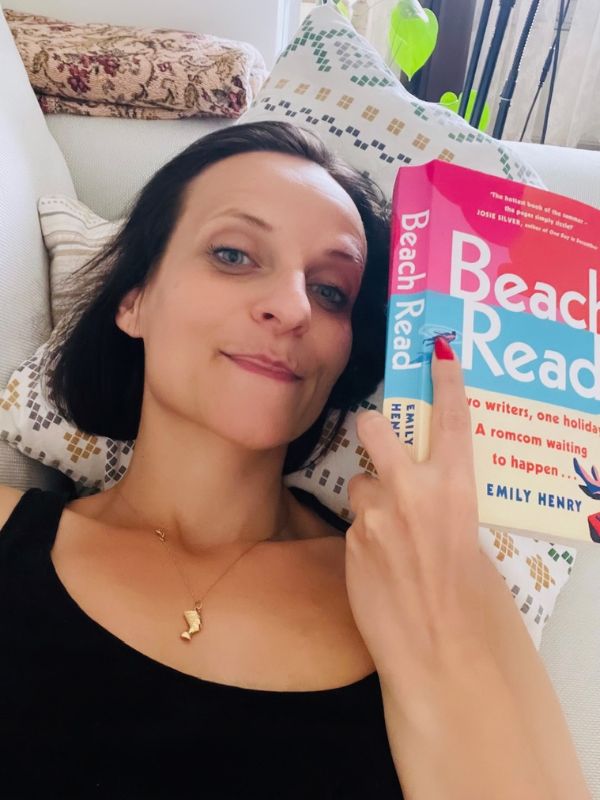 Woman with brunette hair lying on a sofa and holding a book titled "Beach Read"in her left hand.