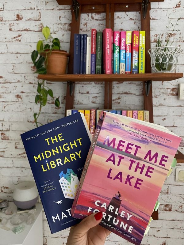 Hand holding two books: the first one is titled "The Midnight Library", the Second book is titled "Meet me At The Lake"; the background is a artistic bookshelf
