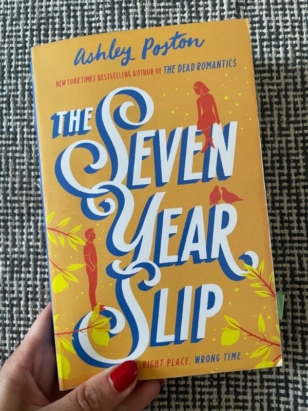 A hand holding an orange-colored book titled: 'The Seven Year Slip' by Ashley Poston, The title's font is big and white, in good contrast  to the orange-yellow background.
