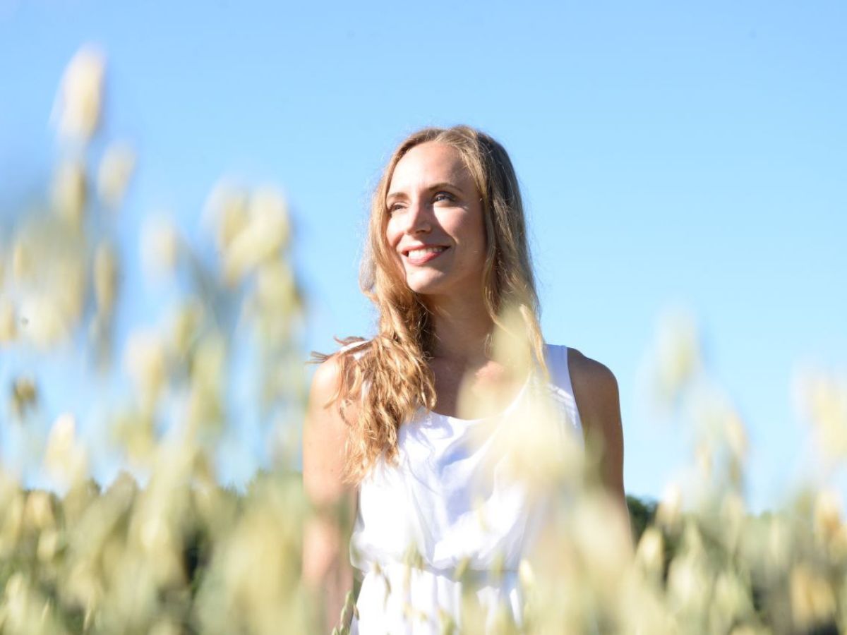 Woman with long blond hair, wearing a white summer dress, stands in a field. The sky is blue.
