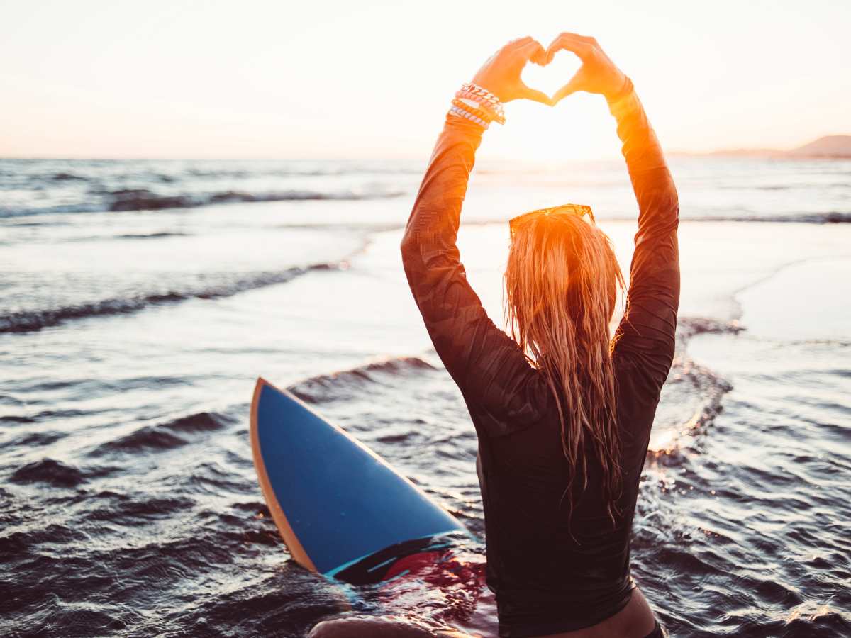 Blond woman is sitting on her surf board, holding up her hand to form a heart with her hands, capturing the sun set.