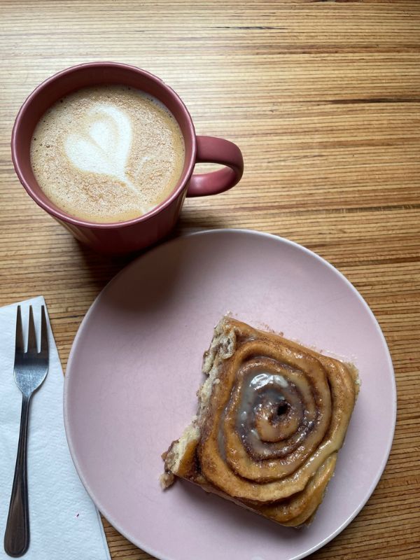 a cup of coffee with a 'milk heart' and on a pink plate a vegan cinnamon roll