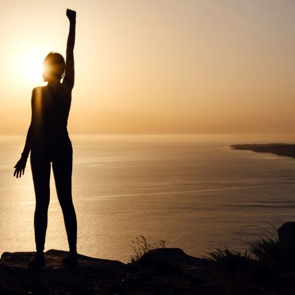 Women standing in front of the sea with a victory pose while watching the sunset 
