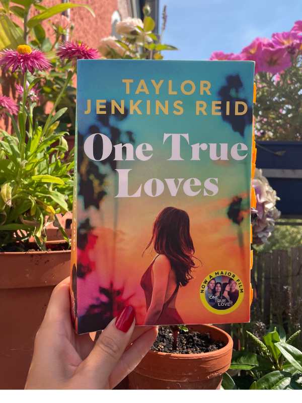 A female hand holding up a book titled: One True Loves. There are pots of flowers in the background. 