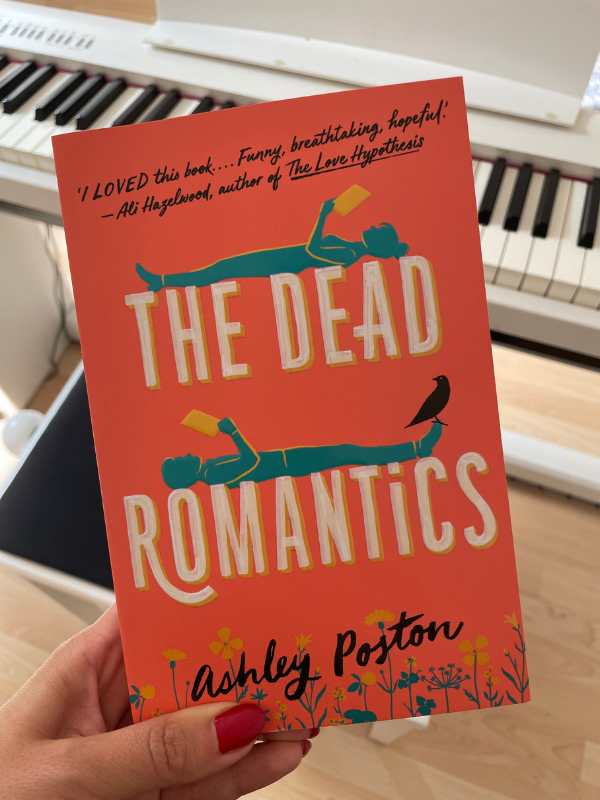 a female hand holding up a book titled The Dead Romantics. There's a white piano in the background.
