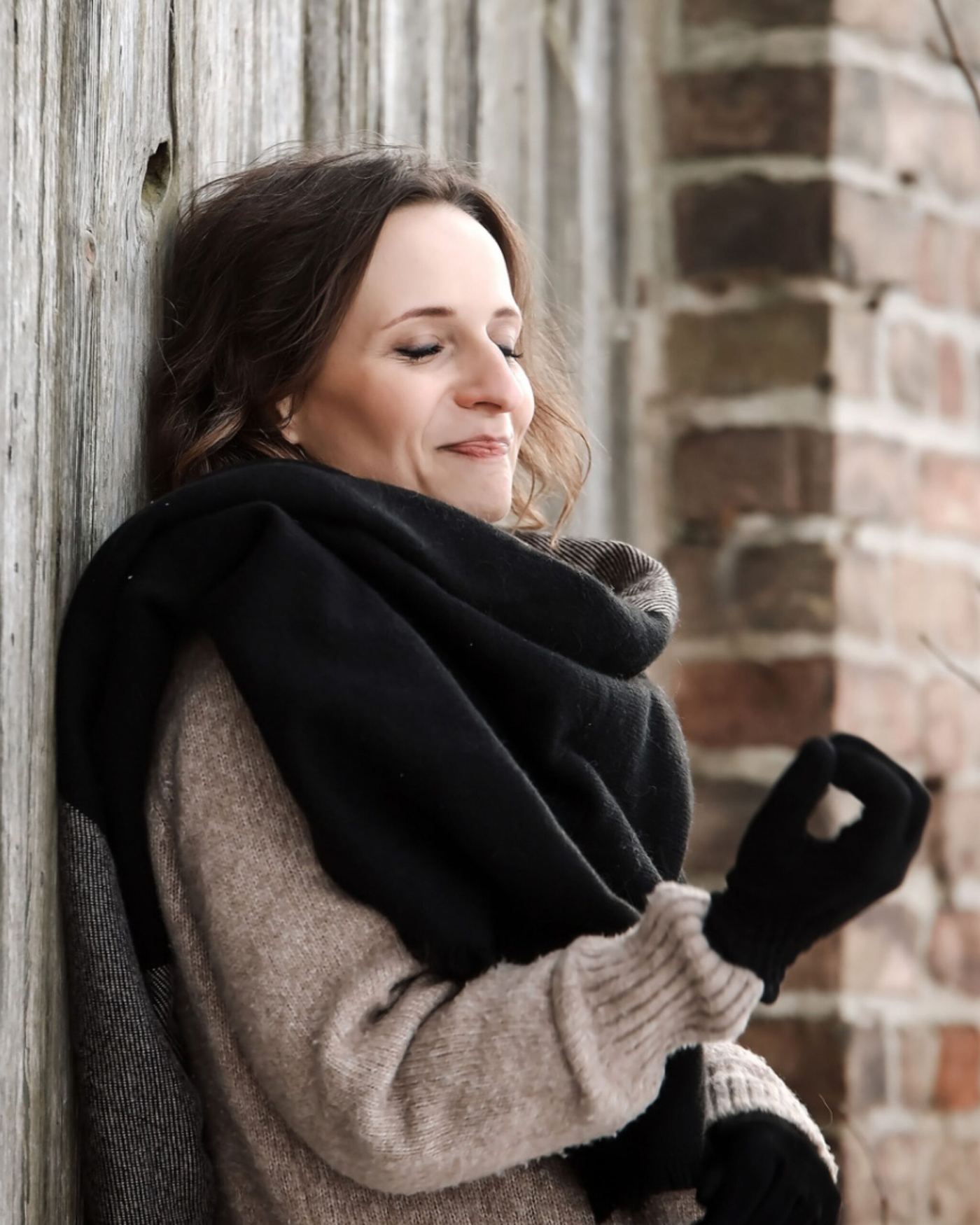 Nadine Kühn with eyes closed and a soft smile, wrapped in a black scarf and leaning against a brick wall in winter