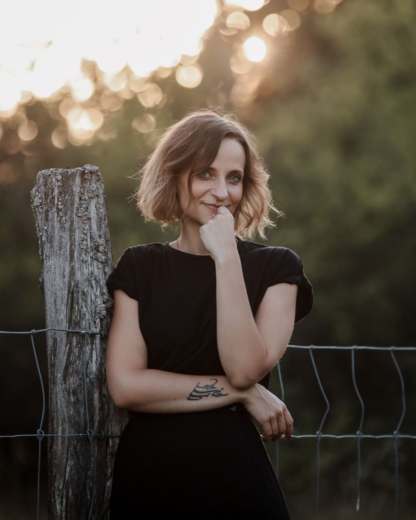 Nadine Kühn leaning against a wooden fence post at golden hour, smiling softly with her hand near her chin