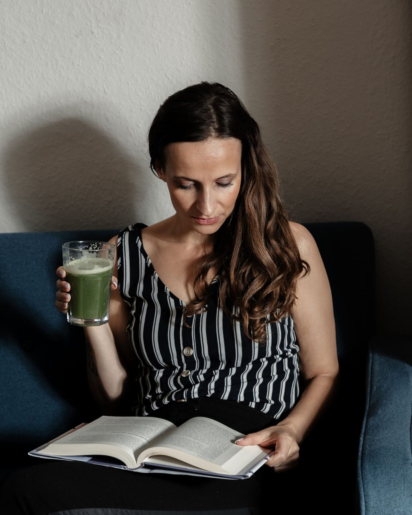 Nadine Kühn reading a book on a blue sofa with a green smoothie in her hand