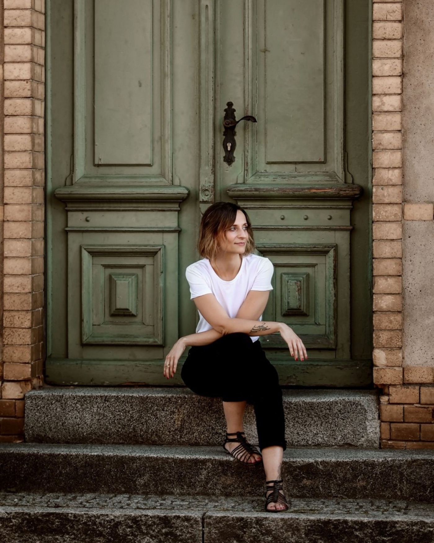 Nadine Kühn sitting on stone steps in front of an old green door, looking over her shoulder with a soft smile
